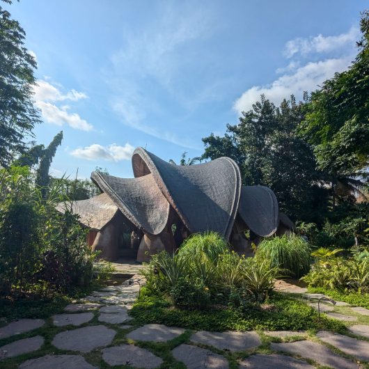A uniquely designed bamboo structure with large curved rooflines sits surrounded by lush vegetation and a stone pathway, under a bright blue sky.