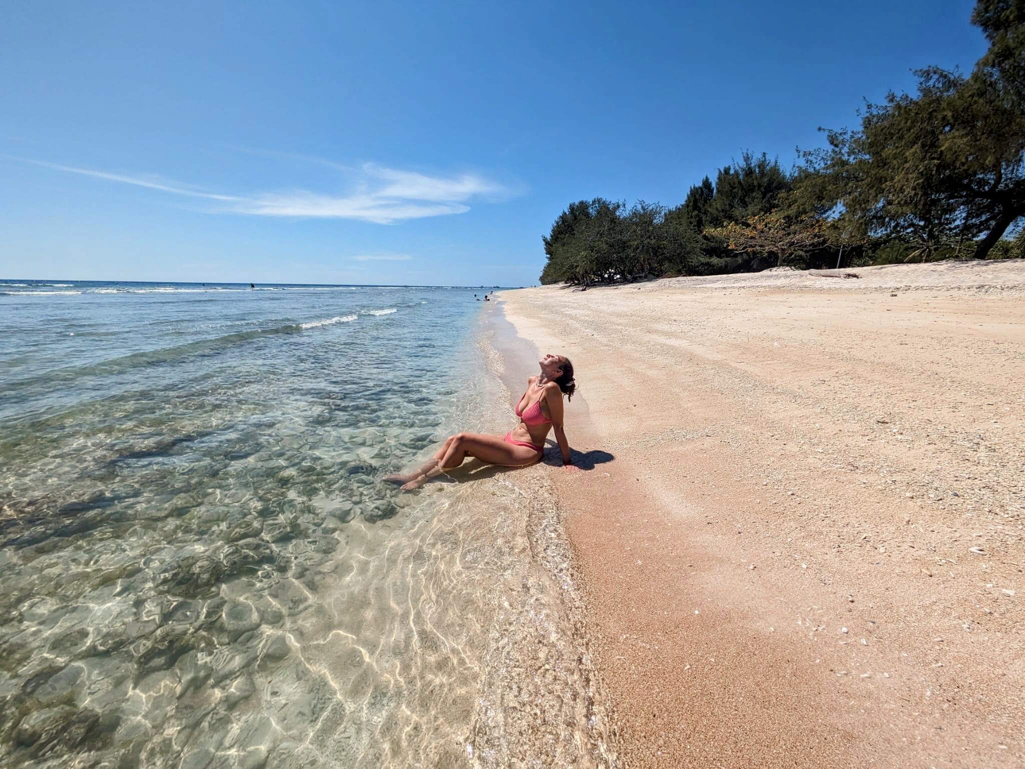 Person in a pink swimsuit relaxes at the edge of a white sand beach in Gili Trawangan with their feet in clear turquoise water, leaning back to enjoy the sun. The quiet shoreline stretches into the distance beneath a bright blue sky, highlighting the calm atmosphere.