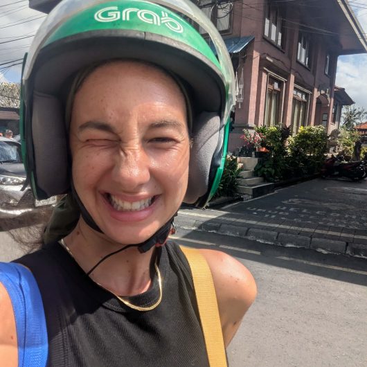 A person is smiling and winking while taking a selfie outdoors, wearing a green Grab motorcycle helmet. They are dressed in a black sleeveless top with a gold necklace and carrying a blue and yellow strap bag, with a street and building visible in the background.