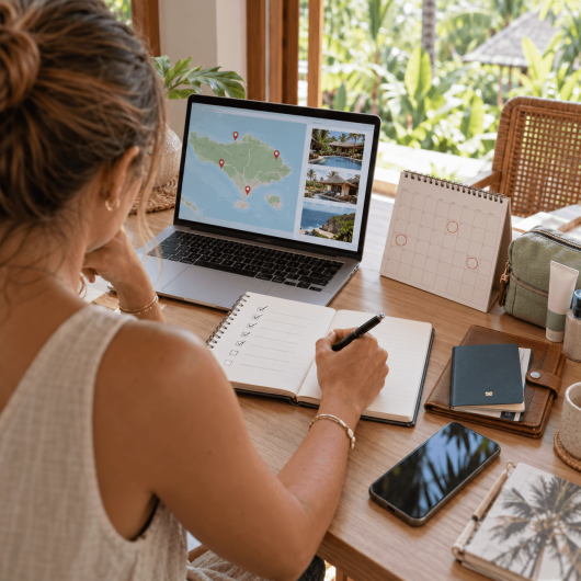 A woman plans a tropical trip at a table with a laptop showing a map and accommodation photos while marking a checklist in a notebook. A calendar passport phone and travel toiletries are arranged beside her as she organizes the details of her upcoming trip.