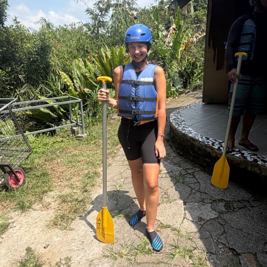 A smiling woman dressed for Ayung River white water rafting wears a blue helmet and life jacket while holding a yellow paddle. Tropical plants surround the outdoor rafting base behind her.