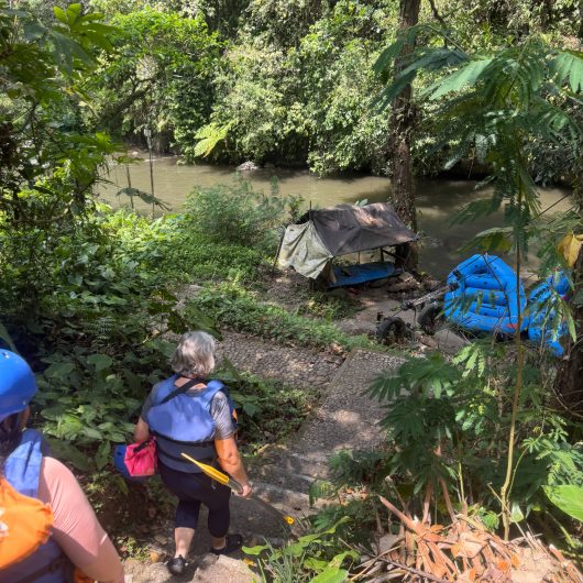 Rafters wearing helmets and life jackets walk down a steep stone path through dense tropical greenery toward blue rafts waiting beside the river.