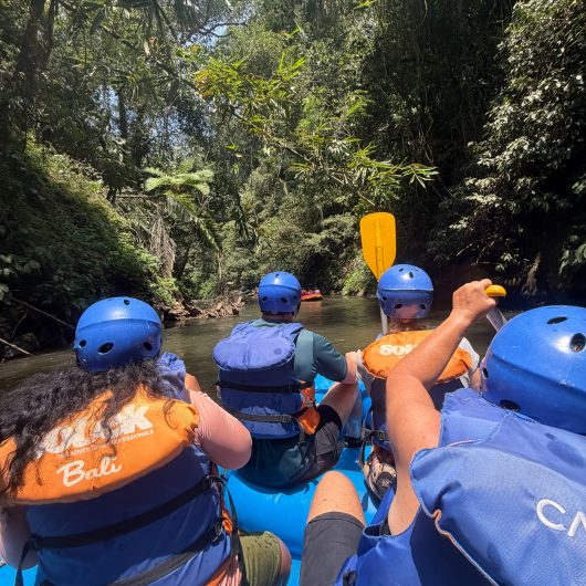 Group of rafters wearing blue helmets and life jackets paddle a blue raft along a narrow stretch of river surrounded by thick tropical jungle. The close view from inside the raft shows what Ayung River white water rafting is like between the faster sections of the river.