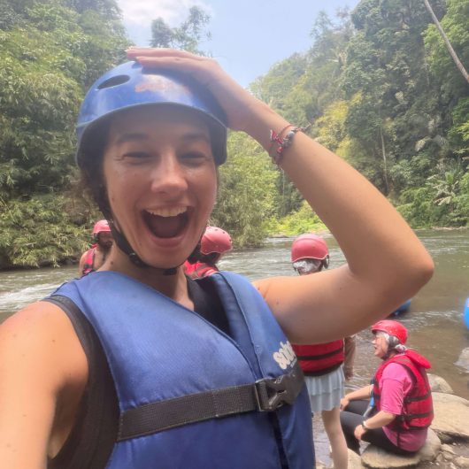 A smiling woman wearing a blue helmet and life jacket takes a selfie beside the Ayung River before white water rafting. Other rafters in red helmets and life jackets wait along the river surrounded by dense tropical forest.