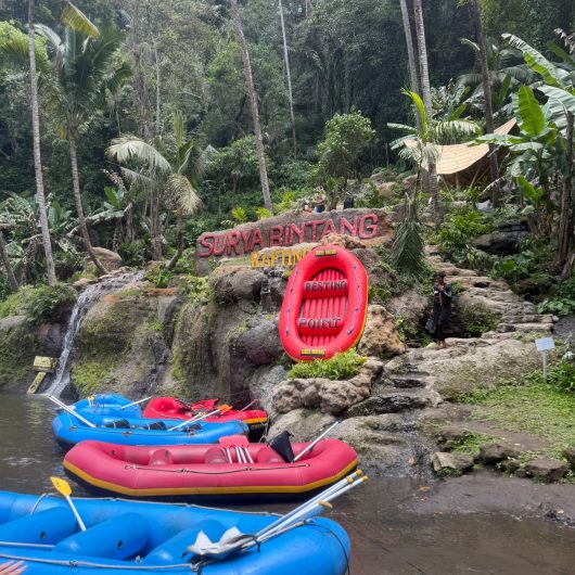 Blue and red rafting boats with paddles rest beside a small waterfall in a lush tropical forest. A large sign on the rocks reads "SURYA BINTANG RAFTING" and a red raft shaped sign reads "RESTING POINT."
