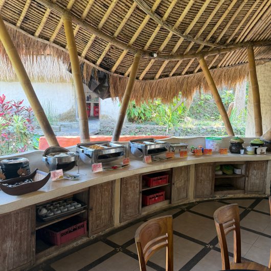 Open air dining area beneath a large thatched bamboo roof with a buffet arranged along a curved counter. Chafing dishes and tableware are set out for rafters to eat after their river trip.