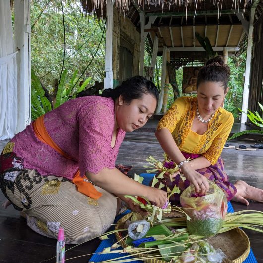 A woman in traditional Balinese clothing teaches another woman how to weave small palm leaf offerings decorated with colorful flowers. They sit together on the floor of an open air pavilion surrounded by tropical greenery.