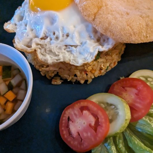 Indonesian fried rice topped with a sunny side up egg and a large crispy cracker. Sliced tomato cucumber and lettuce are served alongside a small bowl of diced vegetables in sauce.