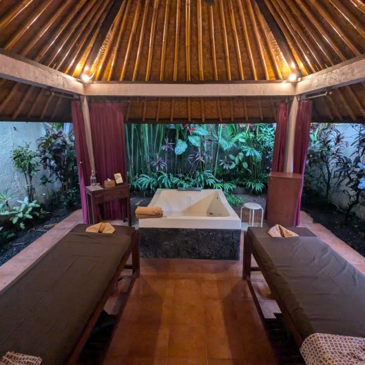 Open air spa treatment room beneath a high bamboo roof with two massage tables and a large white soaking tub. Tropical plants surround the private pavilion and folded towels are arranged for guests.